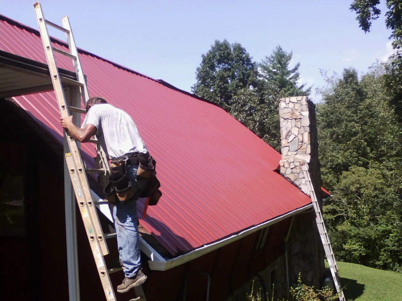 Roofer on a ladder beside a red metal roof and stone chimney — Soffit & Fascia Repair by Roofing Brothers in Pompeys Pillar
